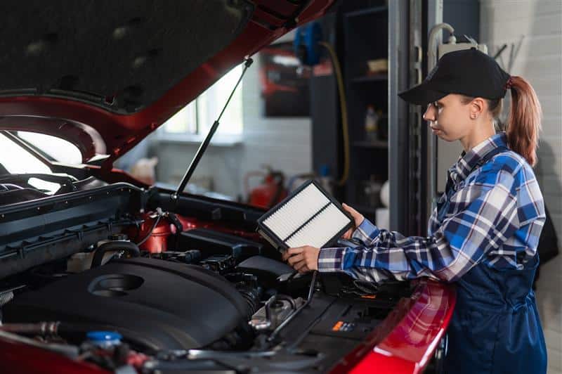 A woman holding an engine air filter next to a car with the bonnet open.